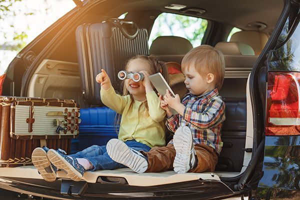 Children in a car trunk with luggage, one using toy binoculars, the other with a tablet, ready for travel.
