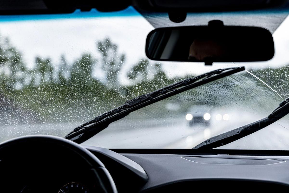 Inside a car during a rainstorm; windshield wipers clearing the glass, road ahead obscured, rear view mirror visible.